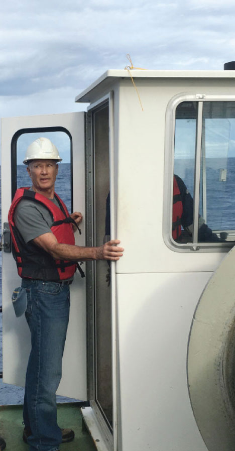 A man, wearing hard hat and life jacket, stands on ship deck near door of a shack on the deck.