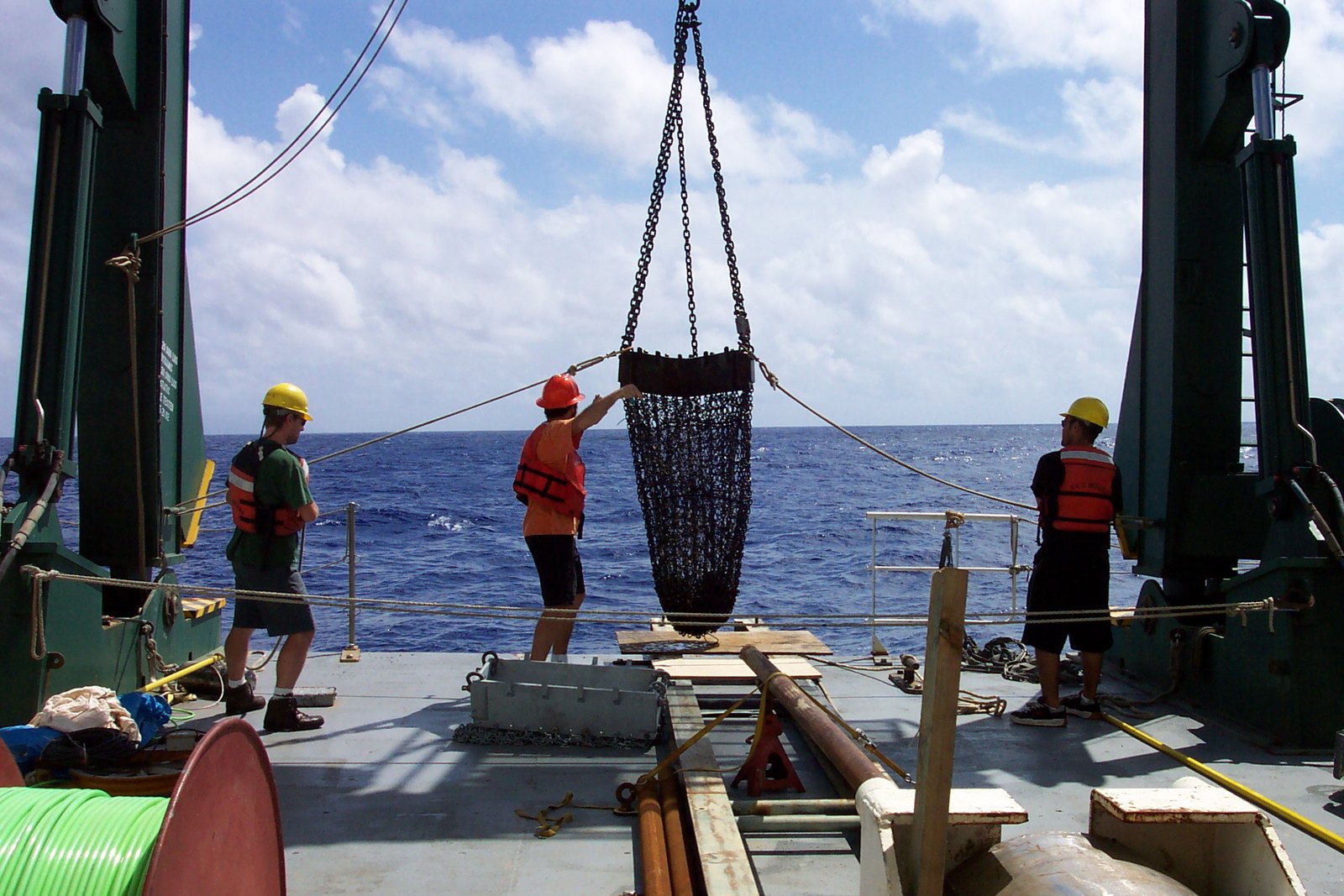 Three people wearing safety gear stand on the stern of a ship to help guide a large ocean-bottom sampler made of chain.