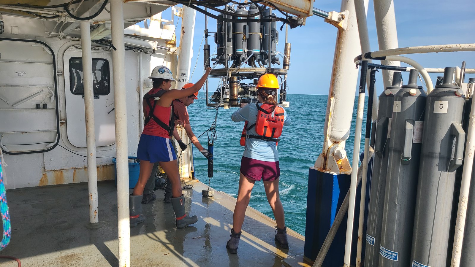 Two scientists aboard a ship hoist a large metal instrument over the waters of the Gulf of Mexico.