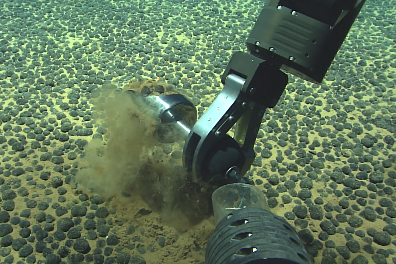 Underwater robotic arm extracting sediment from the ocean floor, with small rounded objects scattered in the background.