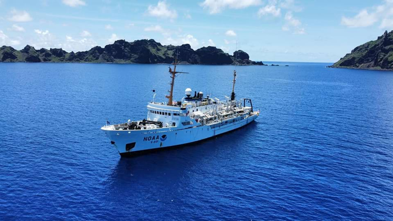 A large white NOAA ship sits at anchor in a calm blue sea, framed by a clear sky with scattered clouds and a distant range of mountains.